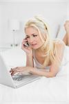 Serious casual young woman using cellphone and laptop in bed at home