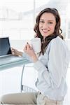 Portrait of a smiling businesswoman with coffee cup in front of laptop in a bright office