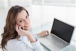 Portrait of a smiling brown haired businesswoman using laptop and cellphone in a bright office