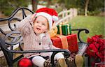 Happy Young Toddler Child Wearing Santa Hat Sitting on Bench with Christmas Gifts Outside.