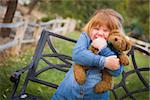 Cute Smiling Young Girl Hugging Her Teddy Bear on Bench Outside.