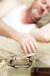 Ashtray with a joint of marijuana burning in the foreground, with a man asleep in the background.  Shallow depth of field.  **Dramatization - no illegal drugs were used in the making of this photograph**