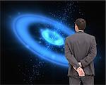 Composite image of businessman standing with hands behind back in front of blue galaxy background