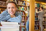 Portrait of a smiling male student with stack of books while others in background at the college library