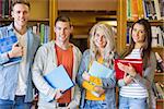 Group portrait of four students with folders standing against bookshelf in the college library