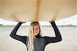 Portrait of a smiling young woman in wet suit holding surfboard over head at beach