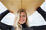 Close up of a smiling young woman in wet suit holding surfboard over head at beach
