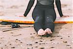 Low section of a young woman in wet suit with surfboard sitting at the beach