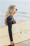 Side view of a beautiful young woman in wet suit holding surfboard at the beach