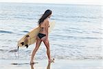 Rear view of a young bikini woman with surfboard walking in water at beach