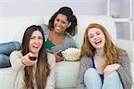 Portrait of cheerful young female friends with remote control and popcorn bowl on sofa at home