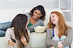 Cheerful young female friends with remote control and popcorn bowl on sofa at home