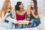 Happy young female friends eating pizza with wine on sofa at home