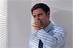 Young businessman peeking through blinds while coughing in office