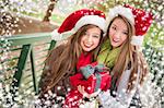 Two Attractive Festive Smiling Mixed Race Women Wearing Christmas Santa Hats Holding a Bow Wrapped Gift Outside with Snow Flakes Border.