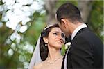 Close-up portrait of bride and groom standing outdoors next to trees in public garden, smiling and looking at each other, in Autumn, Ontario, Canada