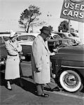 1950s COUPLE IN USED CAR LOT MAN KICKING TIRE