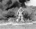 1960s FIREMAN IN ASBESTOS SUIT STANDING IN SMOKE AND FLAMES