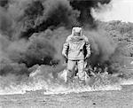 1960s FIREMAN IN ASBESTOS SUIT STANDING IN SMOKE AND FLAMES