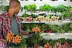 A farm stand with rows of freshly picked vegetables for sale. A man holding bunches of carrots.