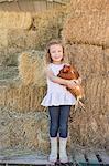 A young girl standing in a hay barn holding a chicken in her arms.