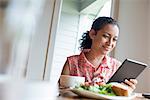 A young woman reading from the screen of a digital tablet, seated at a table. Coffee and a sandwich.