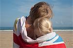 Two children sharing a towel. Back view of a brother and sister sitting on the beach looking out to sea.