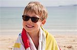 A boy in sunglasses on the beach, with a towel around his shoulders.