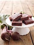 Close-up of beet vegetables next to white cake stand holding beet brownies on wooden background, studio shot