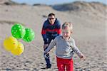 little excited boy running at the beach with colorful balloons and his father waiting