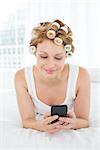 Relaxed young woman in hair curlers text messaging while lying in bed at home