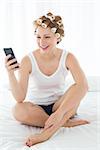 Relaxed young woman in hair curlers text messaging while sitting on bed at home