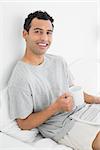 Portrait of a relaxed young man with newspaper and coffee cup sitting on bed at house