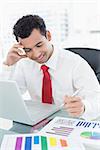 Smiling young businessman with laptop and graphs sitting at office desk