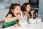 Two smiling young girls eating cereals in the kitchen at home