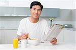 Portrait of a smiling young man having cereals while reading newspaper in the kitchen at home