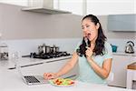 Young woman with laptop eating salad in the kitchen at home
