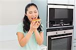 Smiling young woman eating a slice of pizza in the kitchen at home