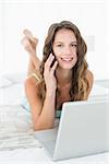 Portrait of a relaxed young woman using mobile phone and laptop in bed at home