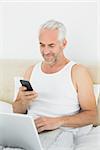 Casual smiling mature man with cellphone and laptop in bed at home