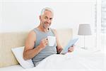 Portrait of a smiling mature man with digital tablet and coffee table sitting in bed at home