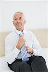 Thoughtful mature businessman adjusting neck tie in bed at home