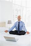 Full length of a mature businessman sitting in lotus pose with laptop on bed at home
