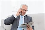 Elegant mature businessman using cellphone while reading newspaper on sofa in living room at home