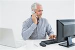 Mature businessman with cellphone, laptop and computer at desk against white background