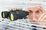 Close-up of a serious mature businessman peeking with binoculars through blinds in the office