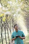 A man standing in an avenue of trees, holding a digital tablet.