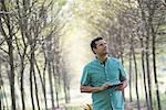 A man standing in an avenue of trees, holding a digital tablet.