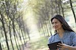 A woman standing in an avenue of trees, holding a digital tablet.