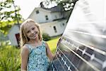 A young girl beside a large solar panel in a farmhouse garden.